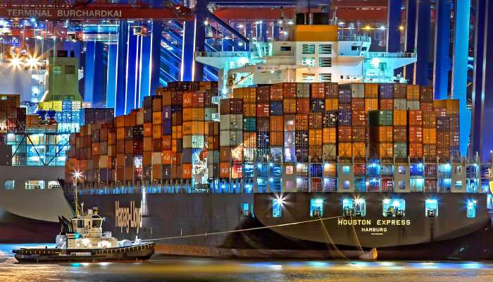 A brightly lit cargo ship at Hamburg harbor with stacked containers and a tugboat.